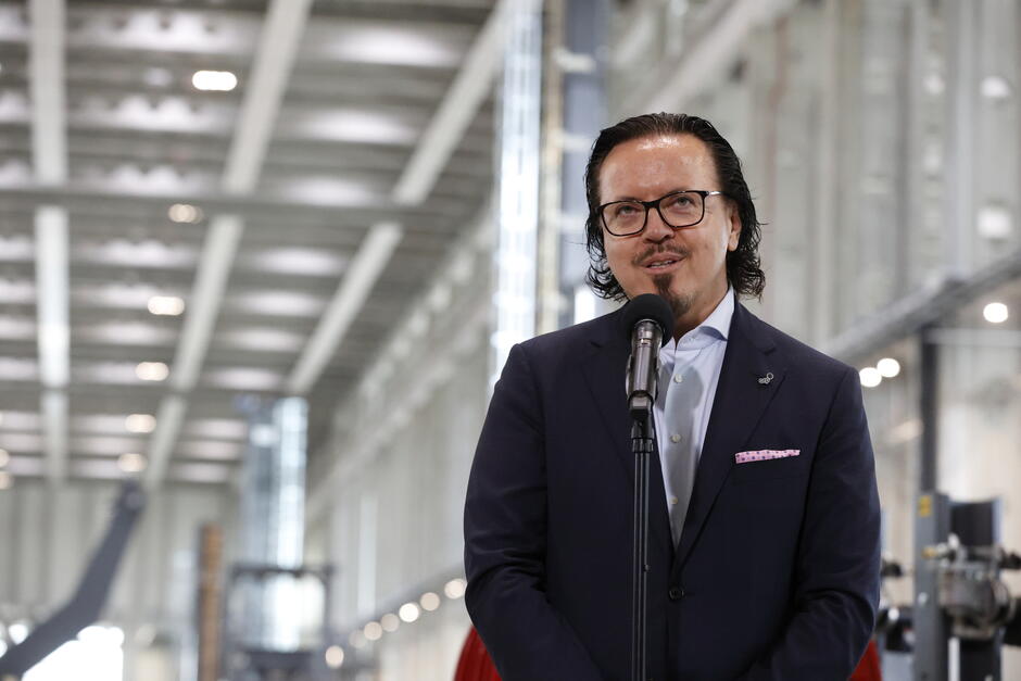 Na zdjęciu widzimy kolejnego mężczyznę przemawiającego do mikrofonu wewnątrz tej samej hali przemysłowej, która była widoczna na wcześniejszych fotografiach z zakładu Baltic Towers.