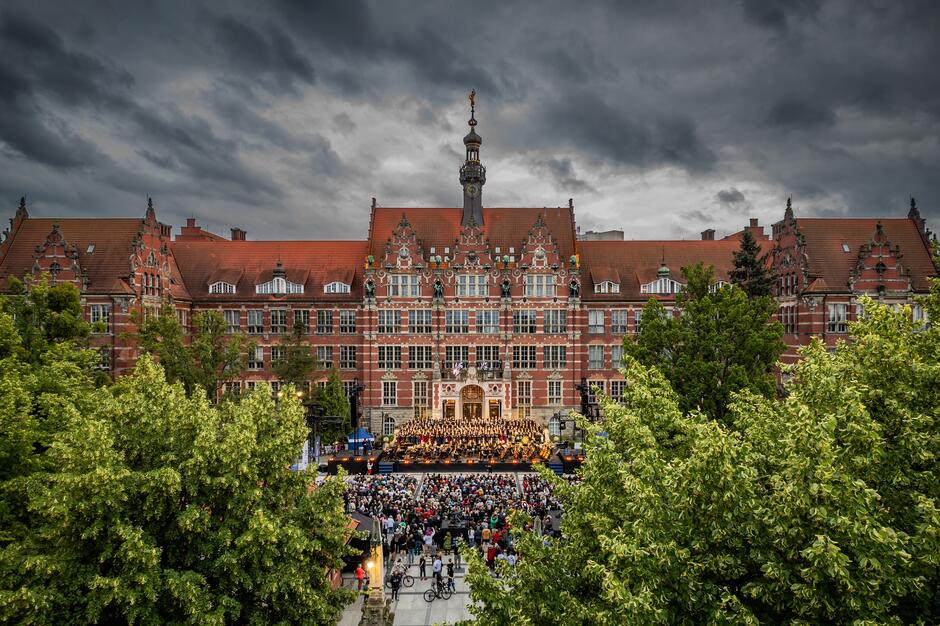 Na zdjęciu widać monumentalny gmach Politechniki Gdańskiej otoczony zielenią, z tłumem ludzi zgromadzonych na koncercie plenerowym przed budynkiem. Na scenie występuje orkiestra i chór, a nad całością unosi się dramatyczne, burzowe niebo, co nadaje wydarzeniu wyjątkową atmosferę
