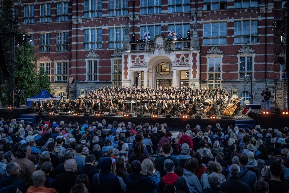 To zbliżenie tej samej sceny pokazuje szczegóły występu orkiestry symfonicznej i chóru, z elegancką fasadą uczelni w tle. Tłum słuchaczy wypełnia plac, wsłuchując się w muzykę w wyjątkowej oprawie architektonicznej