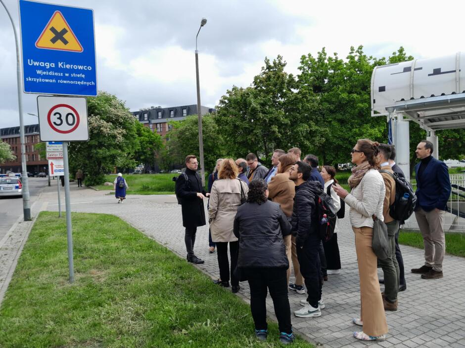Na zdjęciu grupa osób uczestniczy w spotkaniu terenowym w pobliżu miejskiej drogi, gdzie widoczny jest znak informujący o strefie skrzyżowań równorzędnych oraz ograniczeniu prędkości do 30 km/h. Otoczenie sugeruje nowoczesną, zieloną dzielnicę z zabudową miejską w tle i dobrze rozwiniętą infrastrukturą pieszą.
