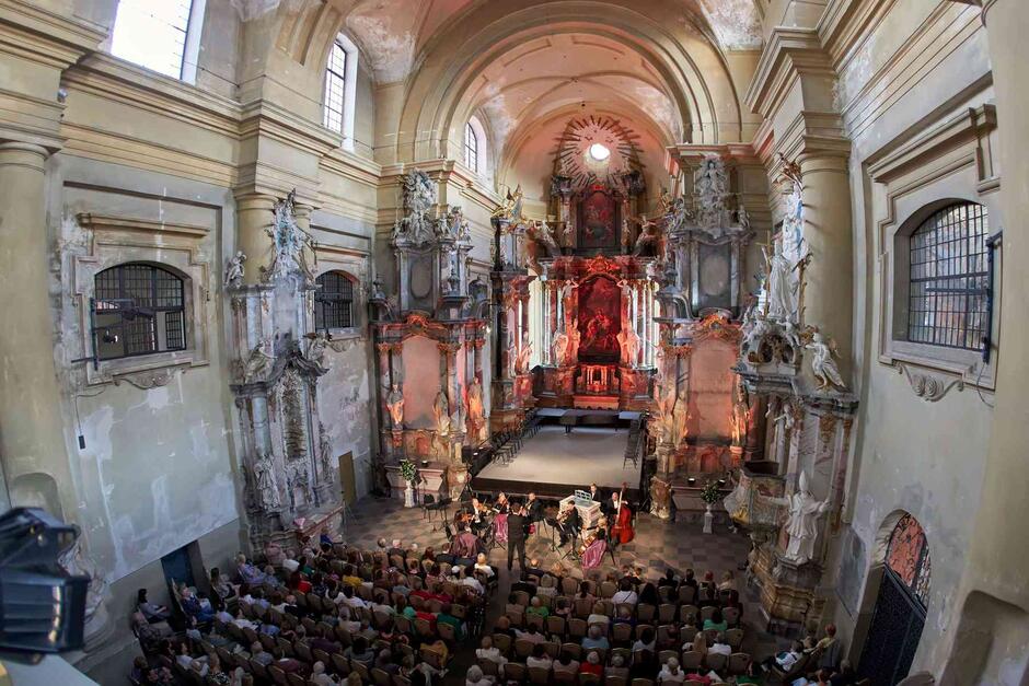 Wnętrze kościoła barokowego widziane z góry. Przed ołtarzem stoi zespół muzyczny, w ławach siedzi publiczność.