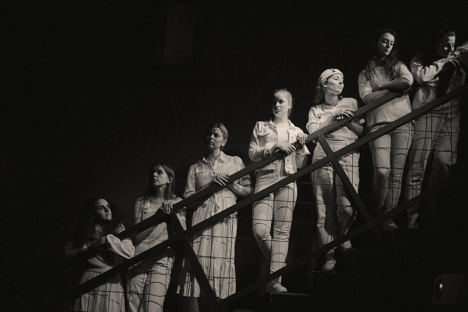 Grupa kobiet stoi na schodach, patrząc w różnych kierunkach, pogrążona w zadumie lub skupieniu, przy przytłumionym świetle. Kompozycja i monochromatyczne kolory nadają zdjęciu teatralną, dramatyczną atmosferę.