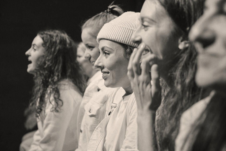 Kobiety siedzą w rzędzie, patrząc w jednym kierunku i reagując emocjonalnie – śmiechem, uśmiechem, ekscytacją. Bliskość ujęcia pozwala dostrzec indywidualne reakcje i buduje poczucie wspólnoty.