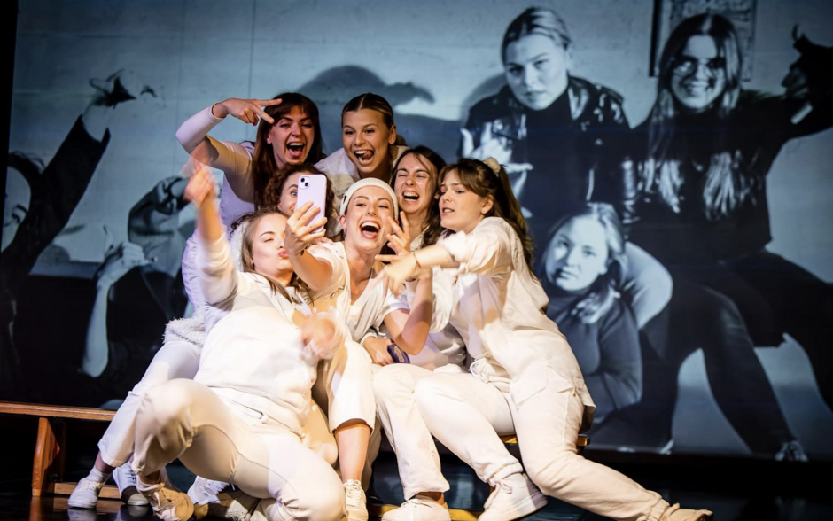 Grupa kobiet robi sobie selfie, śmiejąc się i wygłupiając, co kontrastuje z czarno-białym zdjęciem w tle przedstawiającym te same osoby w bardziej poważnym nastroju. To kolorowe i dynamiczne ujęcie ukazuje radość, energię i wspólną zabawę.