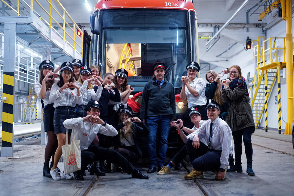 Grupa młodych ludzi w przebraniach policjantów stoi przed tramwajem w zajezdni tramwajowej i pozuje do zdjęcia. Ludzie śmieją się i pokazują serduszka z palców.