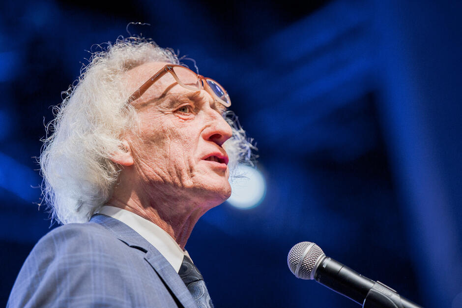 Zdjęcie ukazuje starszego mężczyznę w garniturze, przemawiającego do mikrofonu na scenie, w intensywnym, niebieskim oświetleniu; ma charakter poważny i uroczysty, sugerujący wystąpienie podczas ważnego wydarzenia kulturalnego.