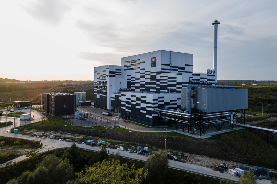 Na zdjęciu widoczna jest nowoczesna spalarnia odpadów o charakterystycznej, geometrycznej elewacji w biało-czarne pasy, z wysokim kominem oraz instalacjami technologicznymi. Otoczona jest infrastrukturą pomocniczą i drogami serwisowymi, a całość znajduje się na obrzeżach miasta, wśród zielonych terenów.