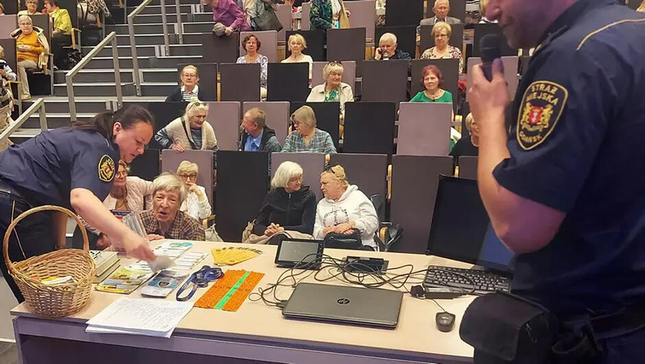W sali wykładowej przy stoliku stoi dwójka strażników miejskich w mundurach, przed nimi siedzi tłum seniorów.