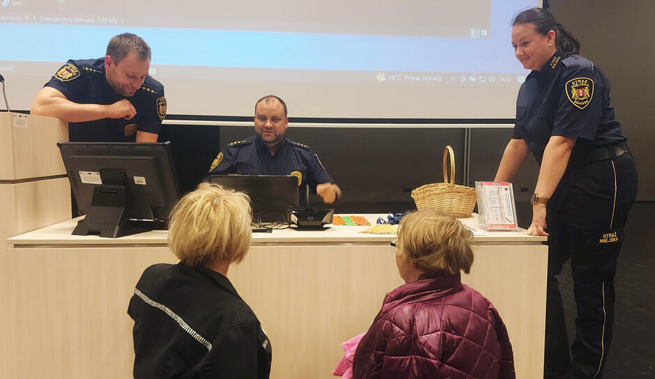 W sali wykładowej na podium siedzi jeden mężczyzna, obok stoi drugi mężczyzna i kobieta. Są funkcjonariuszami Straży Miejskiej w mundurach. Przed nimi stoją dwie osoby tyłem do aparatu.