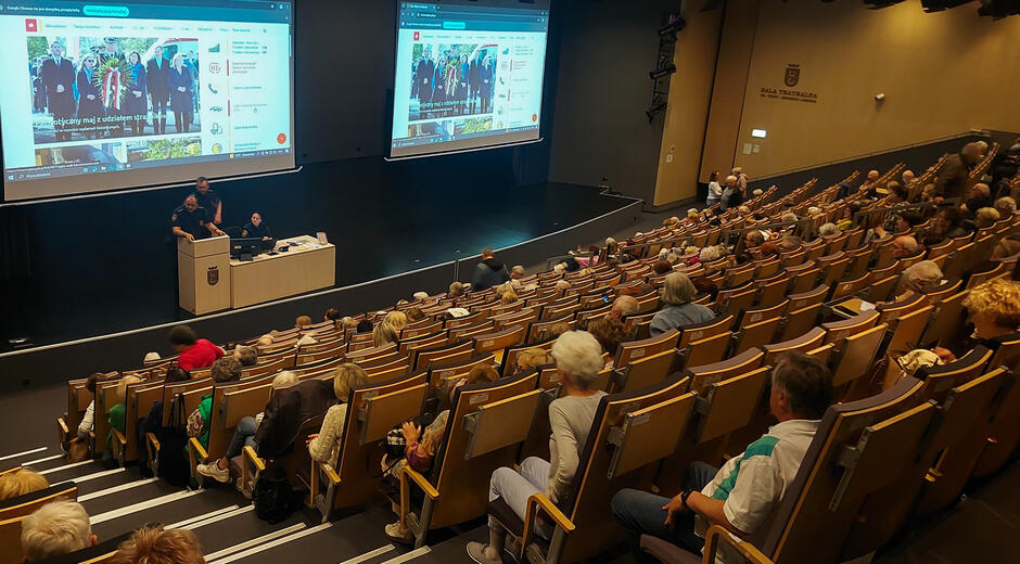 W sali wykładowej siedzi tłum seniorów widzianych od tyłu. Na podium znajduje się trójka strażników miejskich w mundurach, którzy prowadzą wykład. Nad nimi są dwa ekrany z prezentacją.
