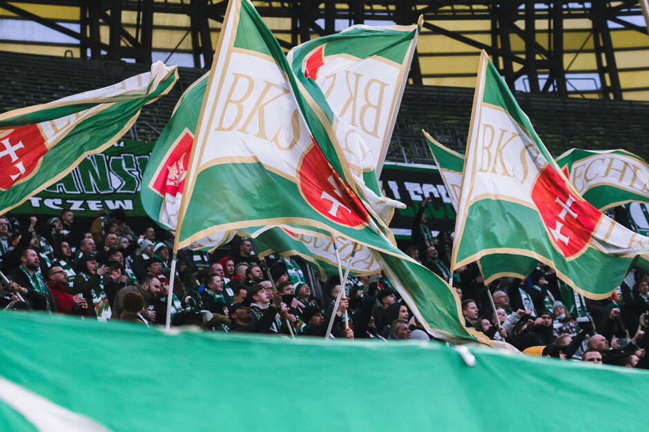 duża grupa ludzi na stadionie, trzymająca zielono-białe flagi z literami BKS na nich. Flagi machają w powietrzu i wydają się machać w tłumie. W tle są rzędy pustych miejsc i transparent z napisem Teczew .
