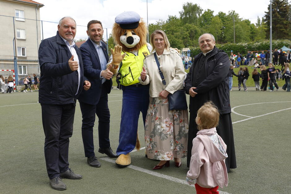 Zdjęcie przedstawia grupę dorosłych wraz z maskotką lwa ubraną w policyjny strój, pozujących wspólnie na boisku sportowym – wszyscy uśmiechają się i pokazują gest kciuk w górę . W tle widać dzieci i rodziców biorących udział w festynie, co tworzy radosną i wspólnotową atmosferę