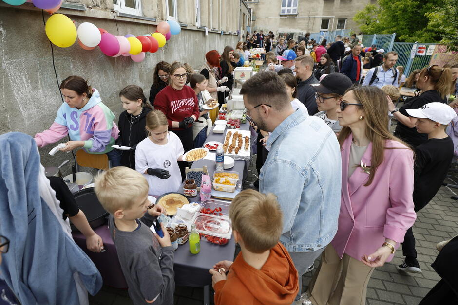 Zdjęcie ukazuje tłum osób przy stoiskach z jedzeniem podczas festynu – dorośli i dzieci kupują oraz serwują gofry, owoce i inne przysmaki. Kolorowe balony i gwar tłumu dodają wydarzeniu energii i rodzinnego charakteru