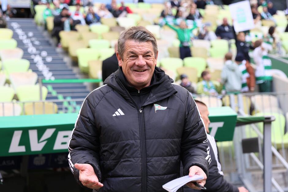 Trener Lechii Gdańsk uśmiecha się szeroko, trzymając notatnik podczas meczu na stadionie w Gdańsku.