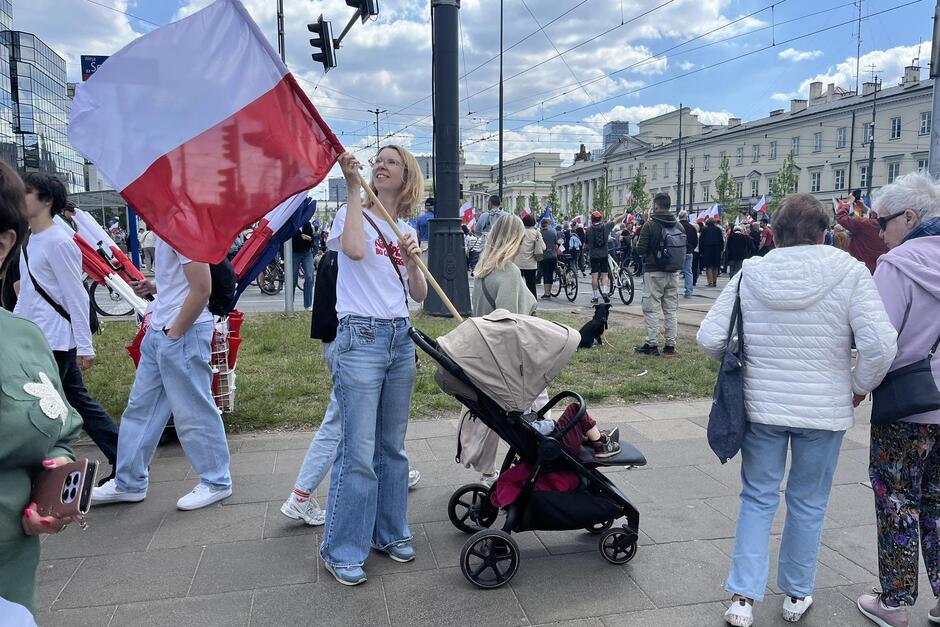 Kobieta stoi na ulicy, pośród tłumu. W jednej ręce trzyma biało-czerwoną flagę, w drugiej - rączkę wózka do wożenia małych dzieci