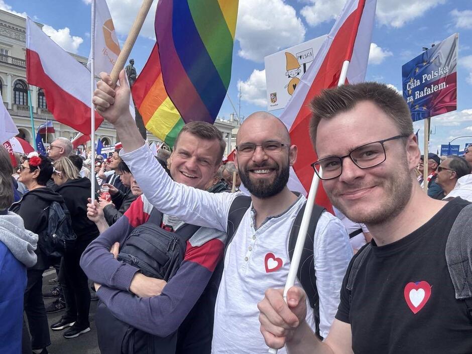 Trzej uśmiechnięci mężczyźni uśmiechają się do zdjęcia, wokół idą inni uczestnicy manifestacji
