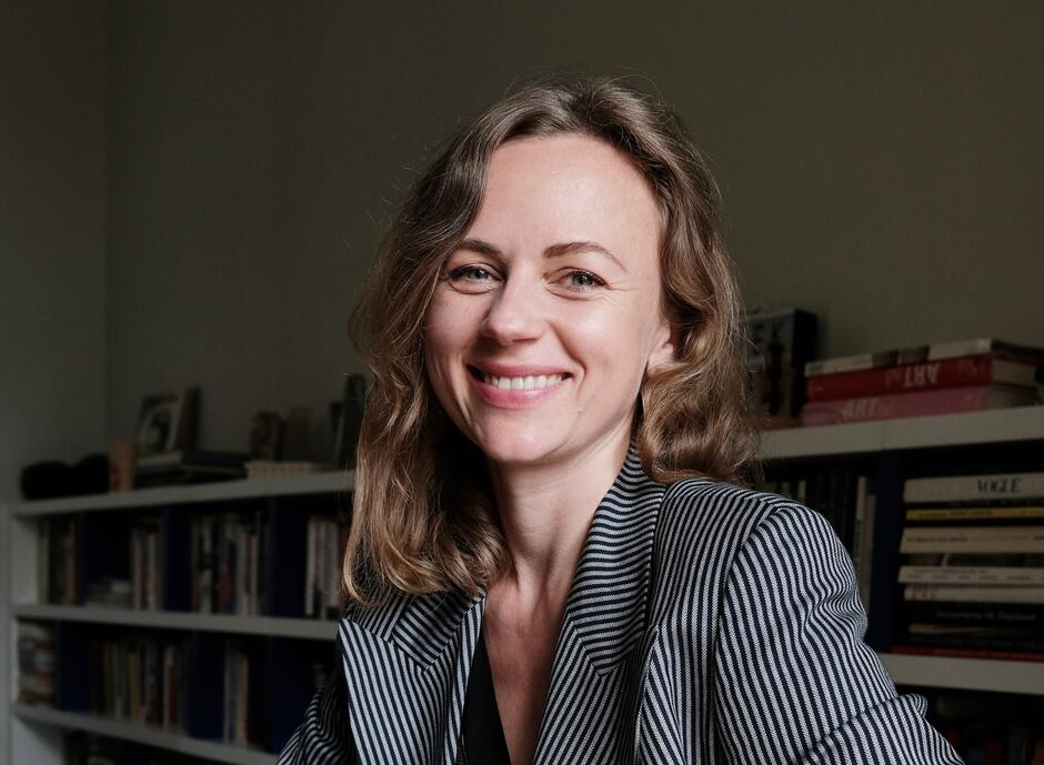 Na zdjęciu kobieta w lekko kręconych włosach, w szarej marynarce. W tle regał z książkami.