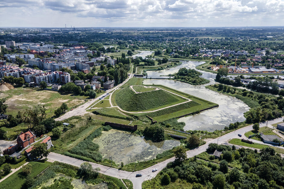 zdjęcie z drona, widać zielony dawny bastion, rzekę, a także zabudowę kamienic w tle