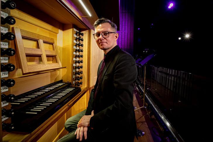 Mężczyzna w okularach siedzi przy dużych organach piszczałkowych, ubrany elegancko w ciemną marynarkę i fioletowy krawat. Otoczenie jest nowoczesne, oświetlone artystycznie fioletowym światłem, co nadaje zdjęciu koncertowego klimatu.