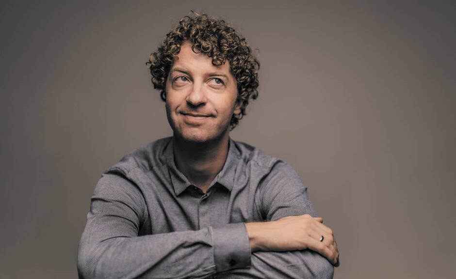 Mężczyzna o kręconych włosach siedzi z założonymi rękami, uśmiechając się lekko i patrząc w bok. Portret utrzymany jest w ciepłym, miękkim świetle, tworząc intymną i przyjazną atmosferę.