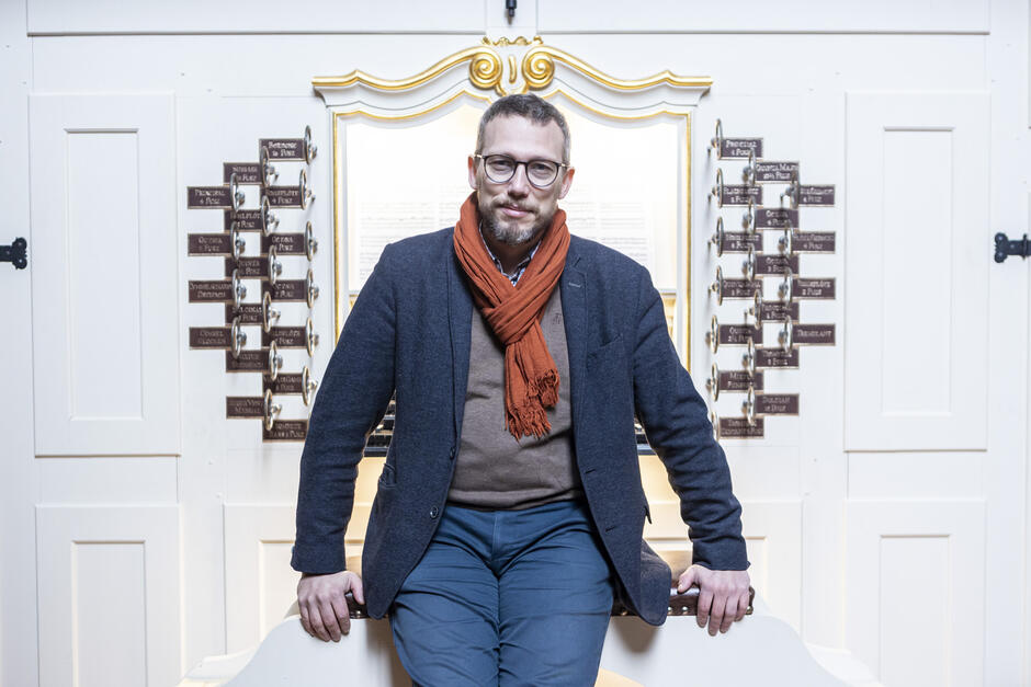Mężczyzna w okularach, ubrany elegancko w ciemną marynarkę i ciepły szalik w kolorze ceglastym, siedzi na ławie przed bogato zdobionym kontuarem organowym. Jasne, barokowe otoczenie instrumentu z licznymi registrami nadaje scenie wrażenie historycznej elegancji i skupienia