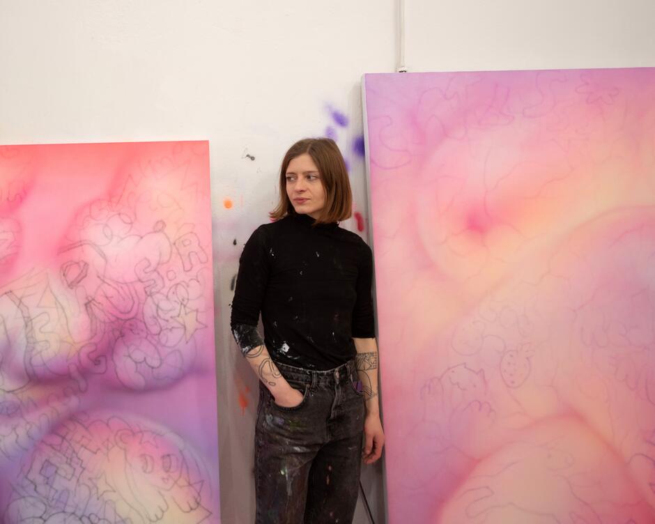 Na zdjęciu kobieta z ręką w kieszeni, w czarnej koszulce. Stoi pomiędzy dwoma obrazami.