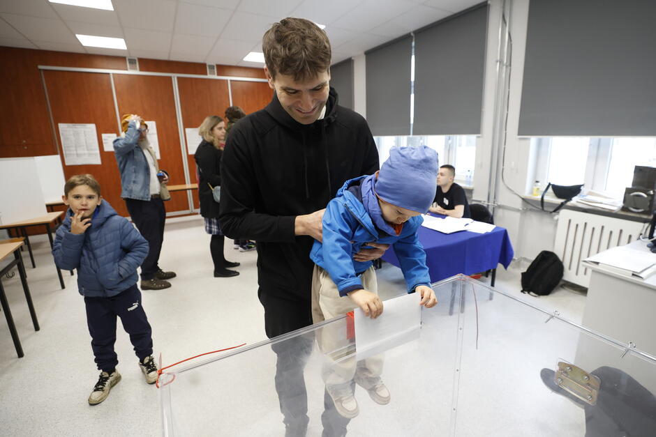 Na zdjęciu widać mężczyznę, który pomaga małemu dziecku wrzucić kartę do przezroczystej urny wyborczej. Mężczyzna uśmiecha się, trzymając dziecko ubrane w niebieską kurtkę i czapkę. Scena rozgrywa się w lokalu wyborczym — w tle widać innych ludzi, w tym dziecko w niebieskiej kurtce oraz osoby czekające w kolejce. Przy stole siedzi członek komisji wyborczej z dokumentami. Atmosfera na zdjęciu jest swobodna i rodzinna, co sugeruje, że udział w wyborach ma charakter obywatelski i międzypokoleniowy.