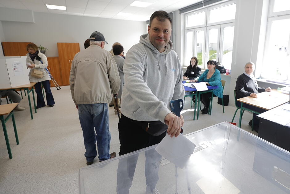 Na drugim zdjęciu widzimy mężczyznę wrzucającego kartę do przezroczystej urny wyborczej. Ubrany jest w jasnoszarą bluzę z kapturem i uśmiecha się w stronę obiektywu. W jednej ręce trzyma laskę, co może wskazywać na ograniczenia ruchowe. W tle widać członków komisji wyborczej siedzących przy stołach oraz innych wyborców oddających głos w kabinach.