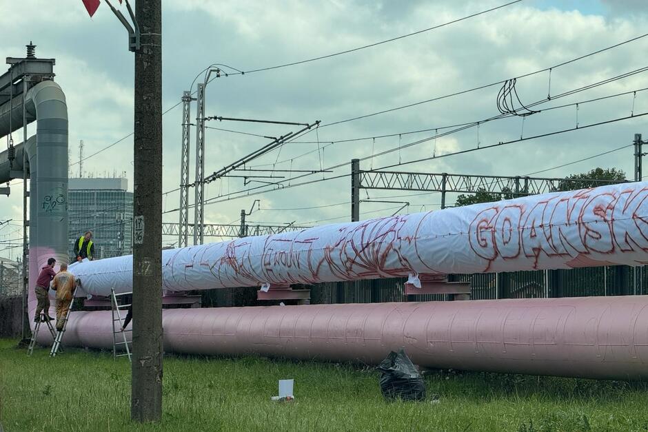 Ciągnące się wzdłuż ulicy rury ciepłownicze, które są już częściowo pokryte jasnoróżową farbą 