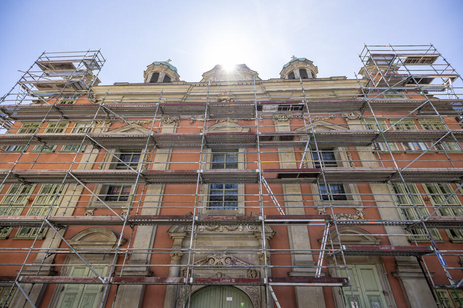 Na zdjęciu widoczna jest zabytkowa fasada budynku pokryta rusztowaniami, co wskazuje na trwające prace renowacyjne. Promienie słońca przebijają się zza kopułowej części dachu, podkreślając historyczny charakter obiektu.
