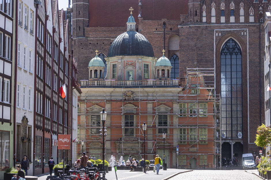 Zdjęcie przedstawia Kaplicę Królewską w Gdańsku, której fasada jest częściowo pokryta rusztowaniami, co świadczy o prowadzonej renowacji. W tle widać monumentalną bazylikę Mariacką, a na pierwszym planie spacerujących ludzi i urokliwą miejską przestrzeń z fontanną. 