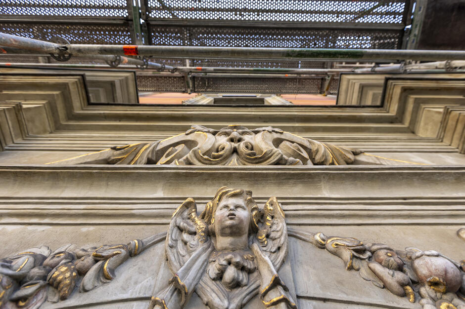 Zdjęcie ukazuje bogato zdobiony detal architektoniczny nad portalem, przedstawiający rzeźbę anioła z rozpostartymi skrzydłami, otoczoną motywami roślinnymi i złoceniami. W tle widoczne są elementy rusztowania, co świadczy o trwających pracach konserwatorskich.
