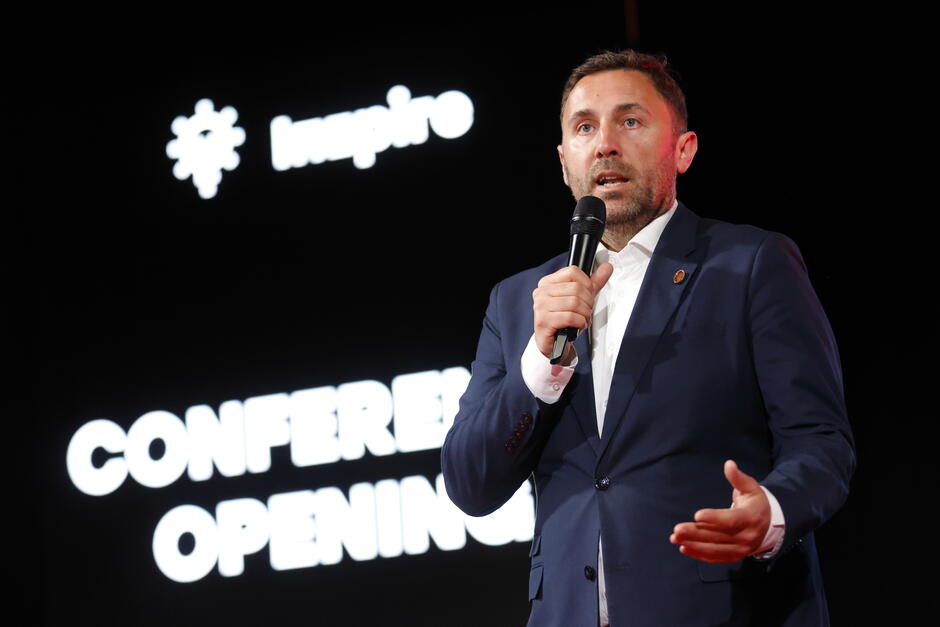 Na zdjęciu widzimy mężczyznę w garniturze przemawiającego do publiczności z mikrofonem w dłoni. Prawdopodobnie jest to jedno z oficjalnych wystąpień otwierających konferencję – co sugeruje widoczny w tle napis „CONFERENCE OPENING” oraz logo i napis „Inspire”. Mężczyzna wygląda na organizatora, gospodarza lub ważnego prelegenta wydarzenia. Scena jest profesjonalnie oświetlona, a tło jest ciemne, co koncentruje uwagę widzów na mówcy.