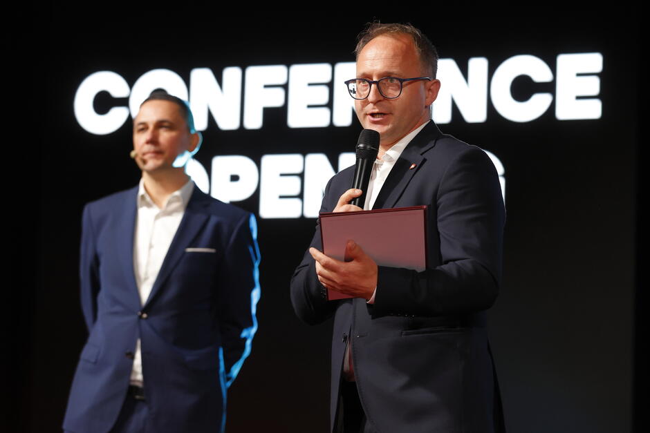 Na zdjęciu widzimy dwóch mężczyzn na scenie podczas oficjalnego otwarcia konferencji — sugeruje to widoczny w tle napis „CONFERENCE OPENING”. Obaj są ubrani elegancko, w garnitury, co podkreśla formalny charakter wydarzenia.