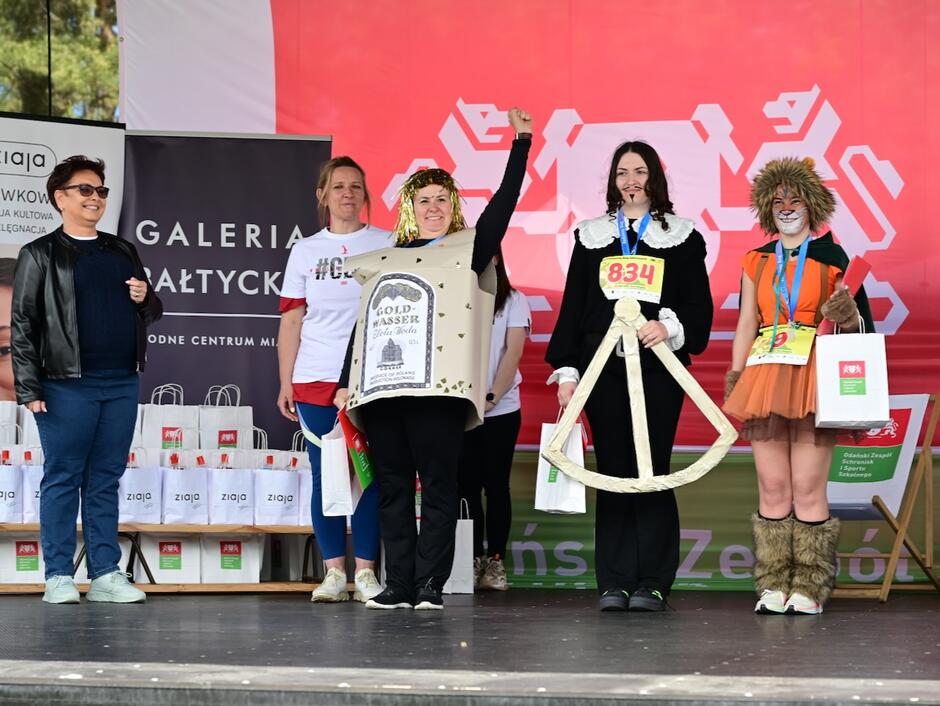 Trzy kobiety w przebraniach inspirowanych Gdańskiem stoją na scenie. Obok dwie kobiety, które prowadzą ceremonie wręczania nagród w konkursie.