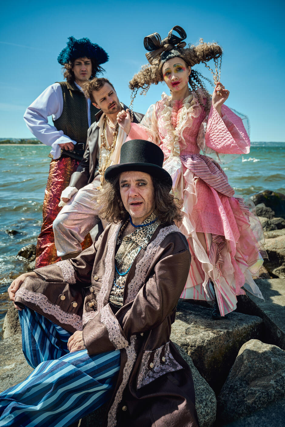 Cztery postacie w teatralnych, fantazyjnych kostiumach pozują na tle morza, stojąc na kamieniach przy brzegu – każda wygląda jak wyrwana z innego snu. Kolorowe stroje, makijaż i stylizacje tworzą surrealistyczną, bajkową atmosferę