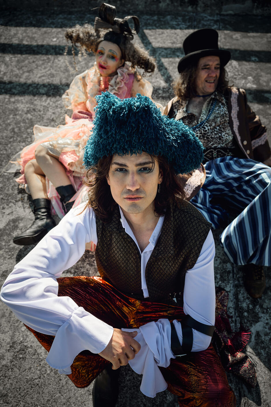 Mężczyzna w niebieskim futrzanym kapeluszu siedzi z przodu, patrząc z powagą w obiektyw, podczas gdy dwie pozostałe postacie siedzą za nim, bardziej rozluźnione. Gra światłem i cieniem dodaje zdjęciu teatralnego napięcia i dramatyzmu