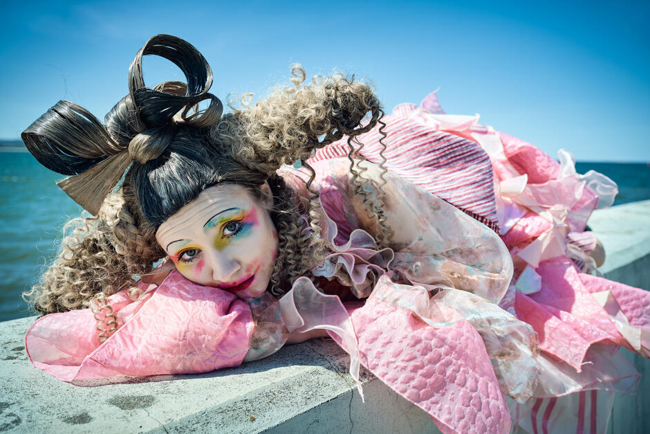 Postać kobieca w cukierkowo-różowej sukni i teatralnym makijażu leży z głową opartą na ramieniu, spoglądając prosto w obiektyw z melancholijnym wyrazem twarzy. Tło morskie i intensywne barwy nadają scenie bajkowy i lekko groteskowy klimat