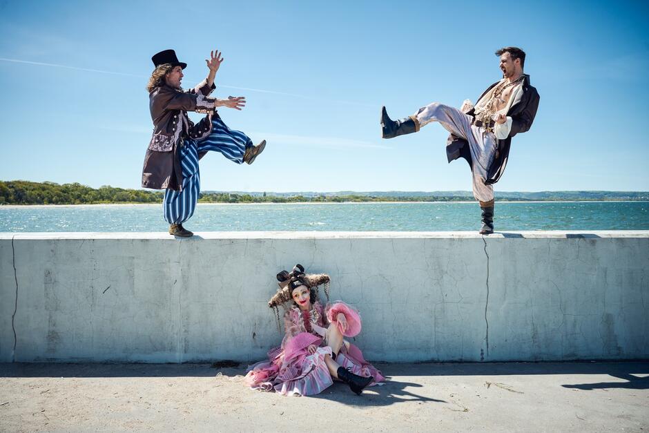 Dwaj mężczyźni w ekstrawaganckich strojach balansują na murku nad wodą, jakby tańczyli lub staczali pojedynek, a kobieta w różowej sukni siedzi na ziemi pod nimi. Kadr pełen ruchu i ekspresji, z nutą absurdu i komizmu