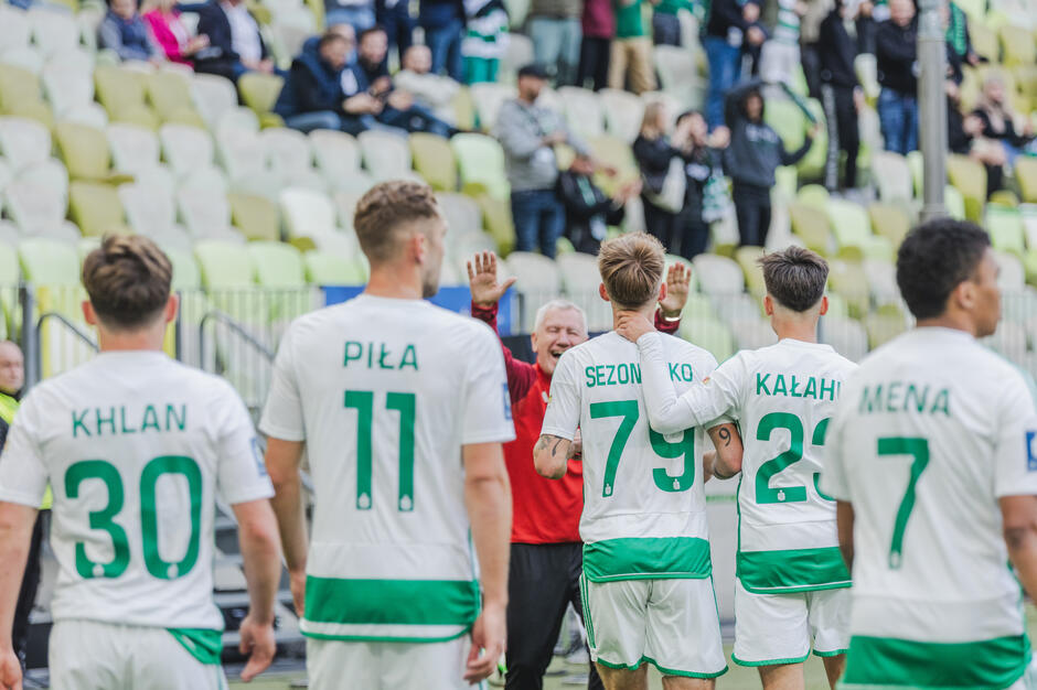  grupa piłkarzy z tej samej drużyny, w białych i zielonych koszulach z numerem 7 na plecach, stoją na boisku z tłumem widzów w tle, gracze są uwięzieni razem w kręgu, z ramionami wokół siebie ramion, w środku znajduje się mężczyzna w czerwonej koszuli, który wydaje się być trenerem lub menedżerem, dając instrukcje graczom, gestyzuje rękami, jakby coś im wyjaśniał.