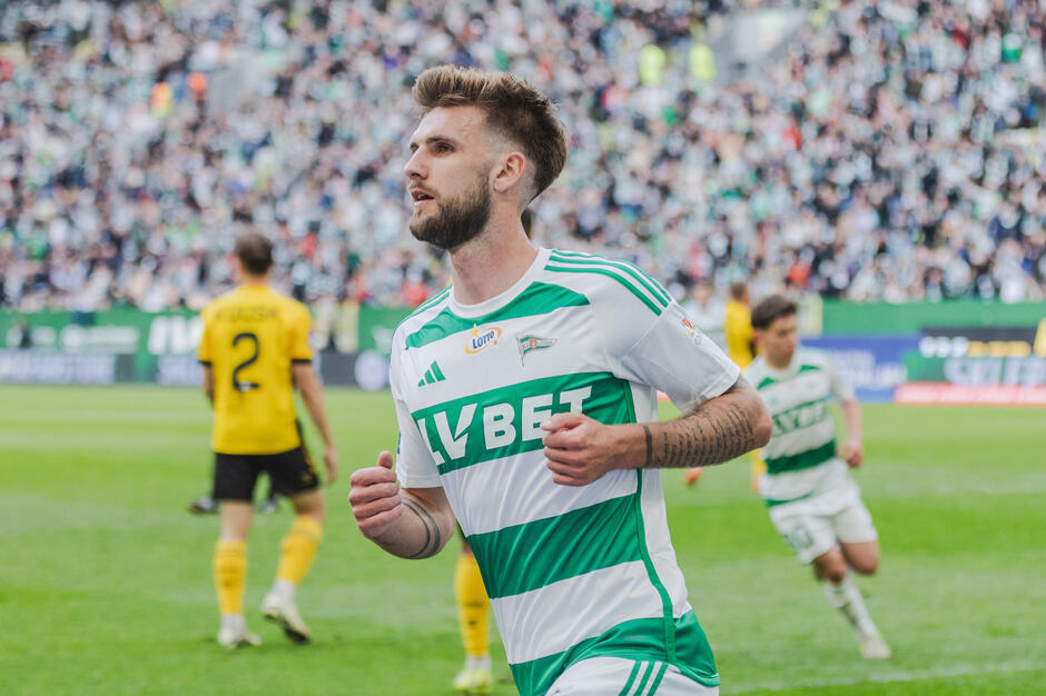  piłkarz na boisku podczas meczu. Ma na sobie zielono-białą koszulkę w pasie z napisem VBET  napisanym na przedniej białymi literami. Gracz ma brodę i patrzy w lewą stronę obrazu z wyraźnym wyrazem na twarzy. W tle są inni gracze z obu drużyn w żółtych koszulach, a na stoiskach widać duży tłum widzów.