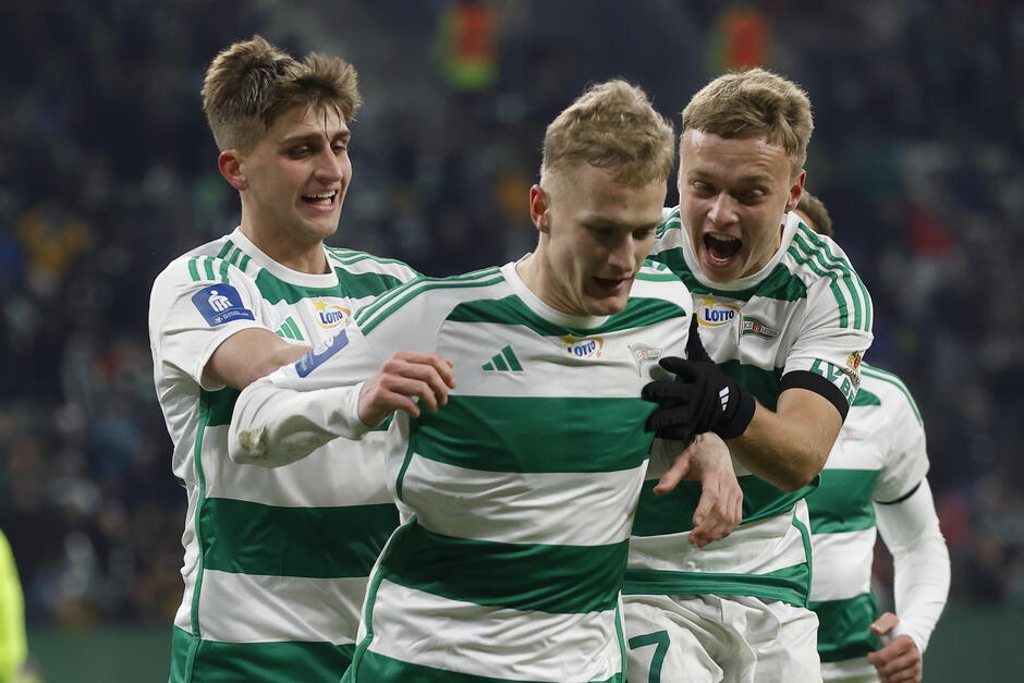 Trzech piłkarzy z tej samej drużyny, ubranych w zielono-białe koszulki pasowe, świętujących gola.Są na boisku, z ramionami wokół siebie i ich twarze są wypełnione radością i emocjami.Gracz w środku, numer 7, jest gratulowany przez swoich kolegów z drużyną, którzy również świętują z nim.Wszyscy trzej gracze uśmiechają się i wydają się być w radości.W tle jest zatłoczony tłum widzów i sędzia.