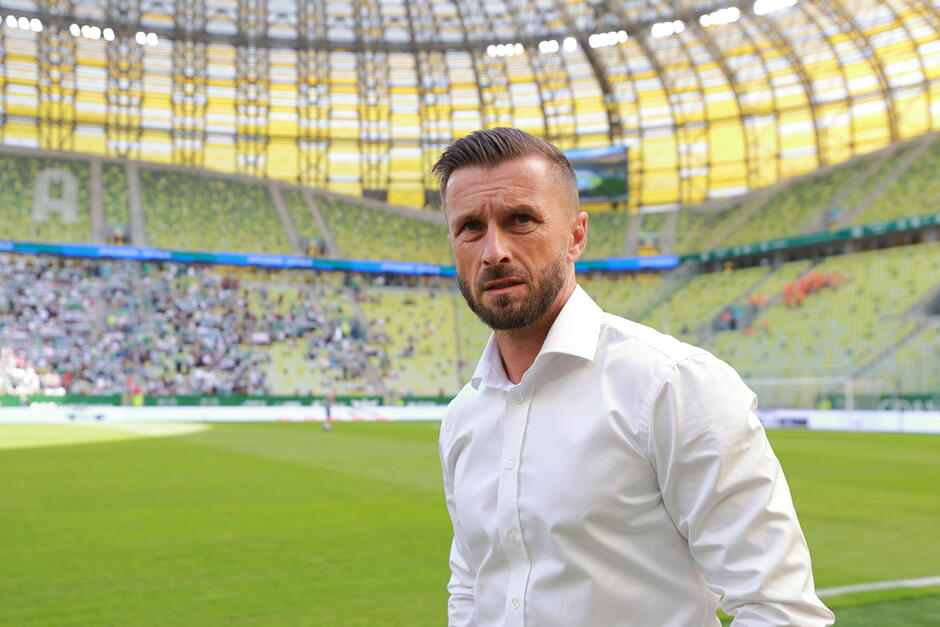  mężczyzna stojący na środku dużego stadionu, nosi białą koszulę i ma poważny wyraz twarzy, stadion jest wypełniony widzami, a stoiska pokryte są zielonymi i żółtymi siedzeniami, w tle jest boisko piłkarskie z kilkoma zawodnikami na boisku, nad człowiekiem widać sufit stadionu z jasnymi światłami świecącymi na nim.