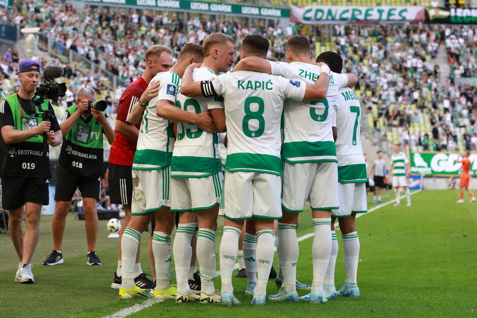  grupa piłkarzy zebranych razem w kręgu na boisku, noszą zielone i białe koszulki z numerami 8 i 7 na nich, gracze stoją blisko siebie, z ramionami wokół swych ramion, w tle jest duży tłum widzów na trybunach, po lewej stronie obrazu można zobaczyć kamerzystę trzymającego kamerę wideo i rejestrującego moment.