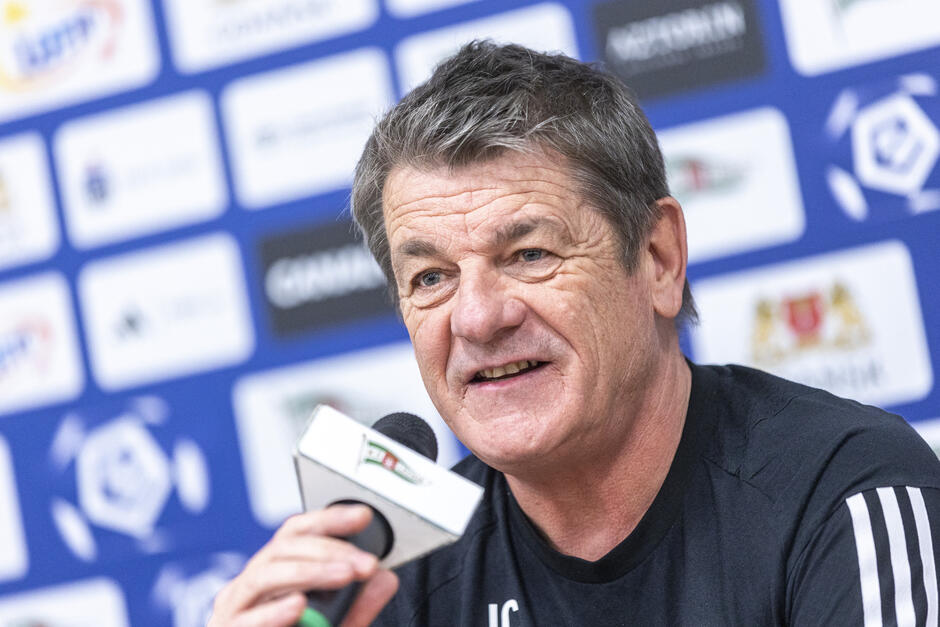  mężczyzna trzymający mikrofon i rozmawiający z mediami, nosi czarną koszulkę z białymi paskami na rękawach i wygląda jak na konferencji prasowej lub wywiadzie, ma krótkie, szare włosy i uśmiecha się szeroko, w tle znajduje się niebieska ściana z wieloma logotypami różnych drużyn sportowych i sponsorów.