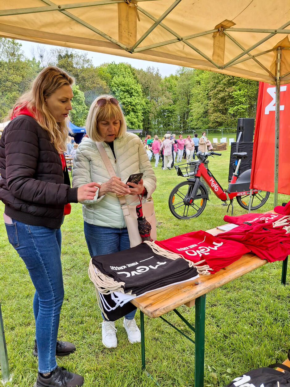 Na zdjęciu dwie kobiety stoją pod namiotem przy stoliku z materiałami promocyjnymi Mevo – jedna z nich tłumaczy coś drugiej, pokazując jej ekran telefonu. W tle widać rower elektryczny Mevo oraz uczestników wydarzenia zgromadzonych na zielonej polanie, co wskazuje na plenerową akcję promocyjną.