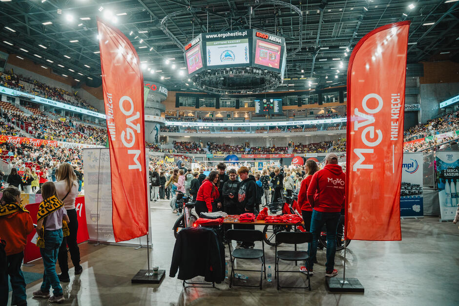 Na zdjęciu widoczna jest strefa promocyjna Mevo zorganizowana wewnątrz ERGO Areny, otoczona licznymi uczestnikami wydarzenia sportowego. Stoisko z czerwonymi materiałami promocyjnymi i dużymi flagami „Mevo – mnie kręci!” przyciąga uwagę odwiedzających, zachęcając do interakcji i poznania systemu rowerów miejskich.
