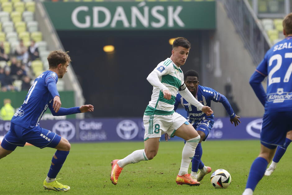  mecz piłki nożnej w toku, zawodnicy są na boisku, a trybuny wypełnione są widzami, na pierwszym planie znajduje się zawodnik z drużyny przeciwnej, ubrany w zielono-białą paskową koszulkę z numerem 18 i drybling piłką w kierunku bramki, ściga go dwóch innych zawodników z obu drużyn, jeden w niebieskiej koszulce, którzy również noszą niebieskie koszulki, zawodnik w zielonej koszuli biegnie do przodu, z wyciągniętymi ramionami i zdecydowanym wyrazem na twarzy, podczas gdy drugi zawodnik próbuje go pokonać. Za nim dwójka zawodników w niebiesko-zielonej koszule biegnie w jego kierunek z wyprostowanymi rękami, próbując zablokować strzał. piłka piłkarska jest widoczna przed nimi i wydaje się być w powietrzu, jakby miała zamiar ją kopnąć.