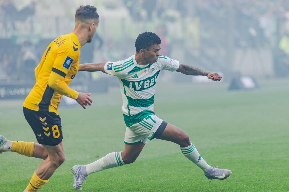  Dwóch piłkarzy w akcji podczas meczu. Gracz po lewej stronie ma na sobie żółtą koszulkę z numerem 8 i biegnie w kierunku prawej strony ma również na głowie tę samą koszulką co gracz w zielono-białej koszulki paskowej. Na pierwszym planie jest gracz z drużyny przeciwnej, w biało-zielonych paskowych spodniach, który odsuwa piłkę od niego. Jego ramiona są wyciągnięte, a jego ciało lekko wygięte do przodu, jakby próbował przejechać obok drugiego gracza. W tle stoiska są wypełnione widzami, i pole jest pokryte mgłą.