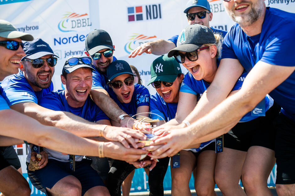 Grupa radosnych osób w niebieskich koszulkach wspólnie trzyma trofeum, świętując zwycięstwo na tle baneru z logo sponsorów.